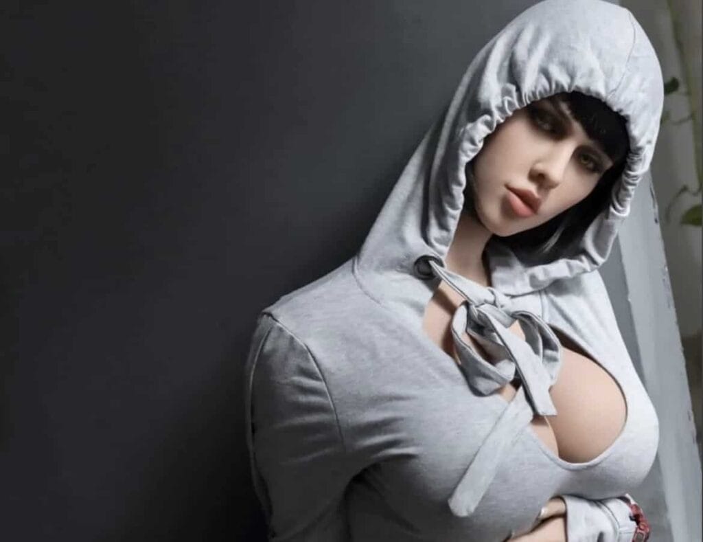 Realistic sex doll wearing a gray hoodie with tied front and hood up, pose on black background.