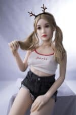 Realistic sex doll with blonde pigtails, wearing a Paris-themed tank top and black denim shorts, posed with deer antler headband.